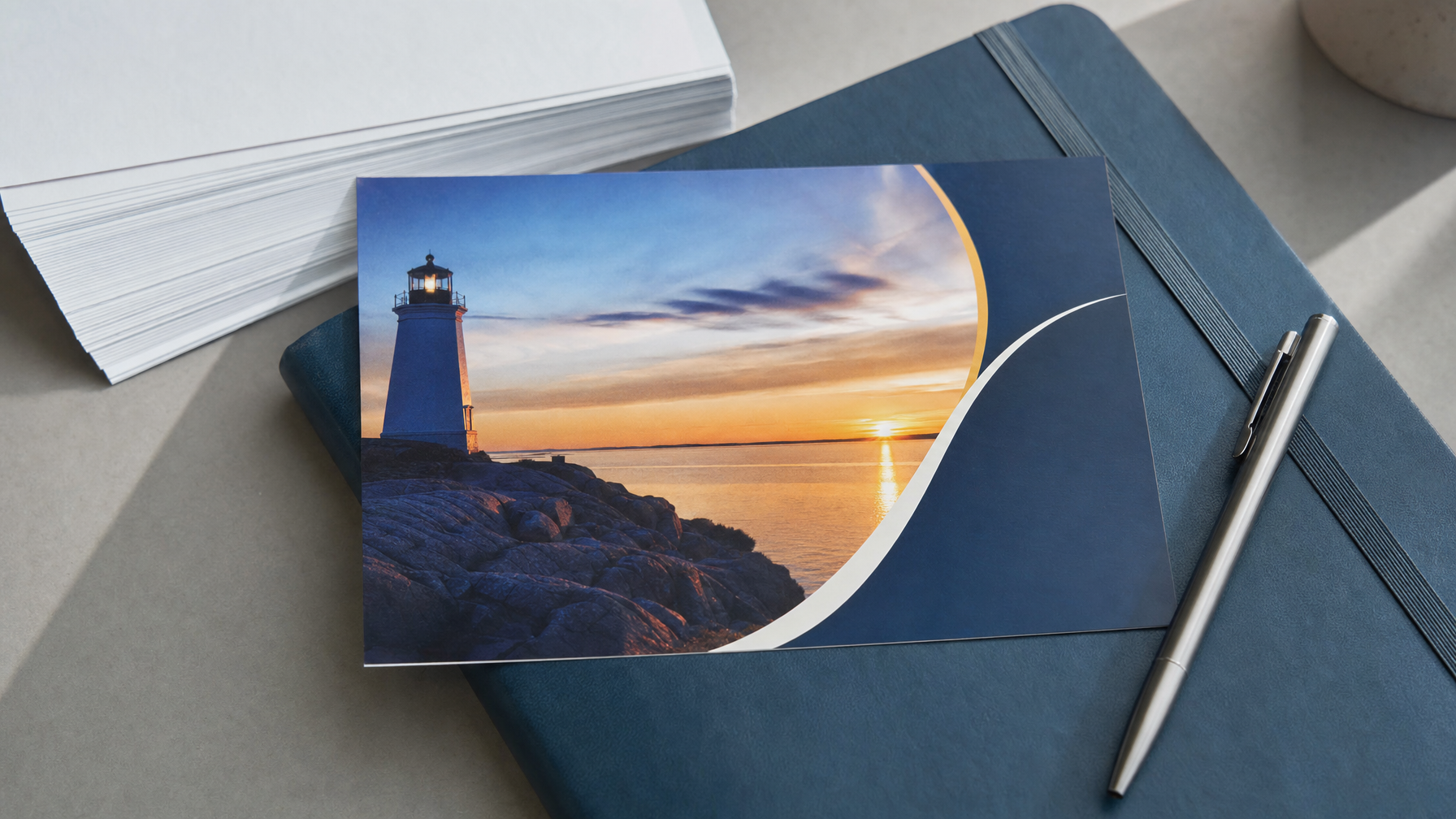 A scenic postcard featuring a lighthouse at sunset rests on a dark notebook beside a pen and a stack of blank cards on a tabletop.