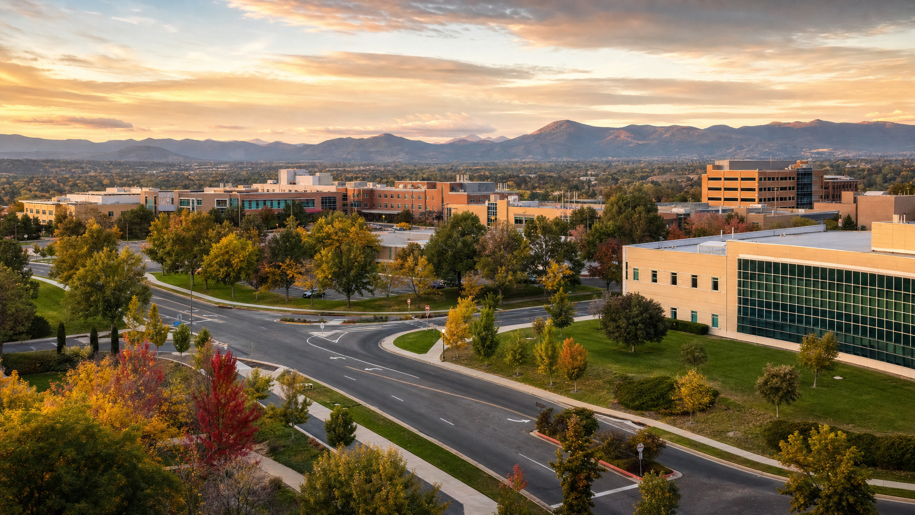 A landscaped business or institutional campus sits below a mountain range at sunset, with tree-lined roads and modern buildings in the foreground.