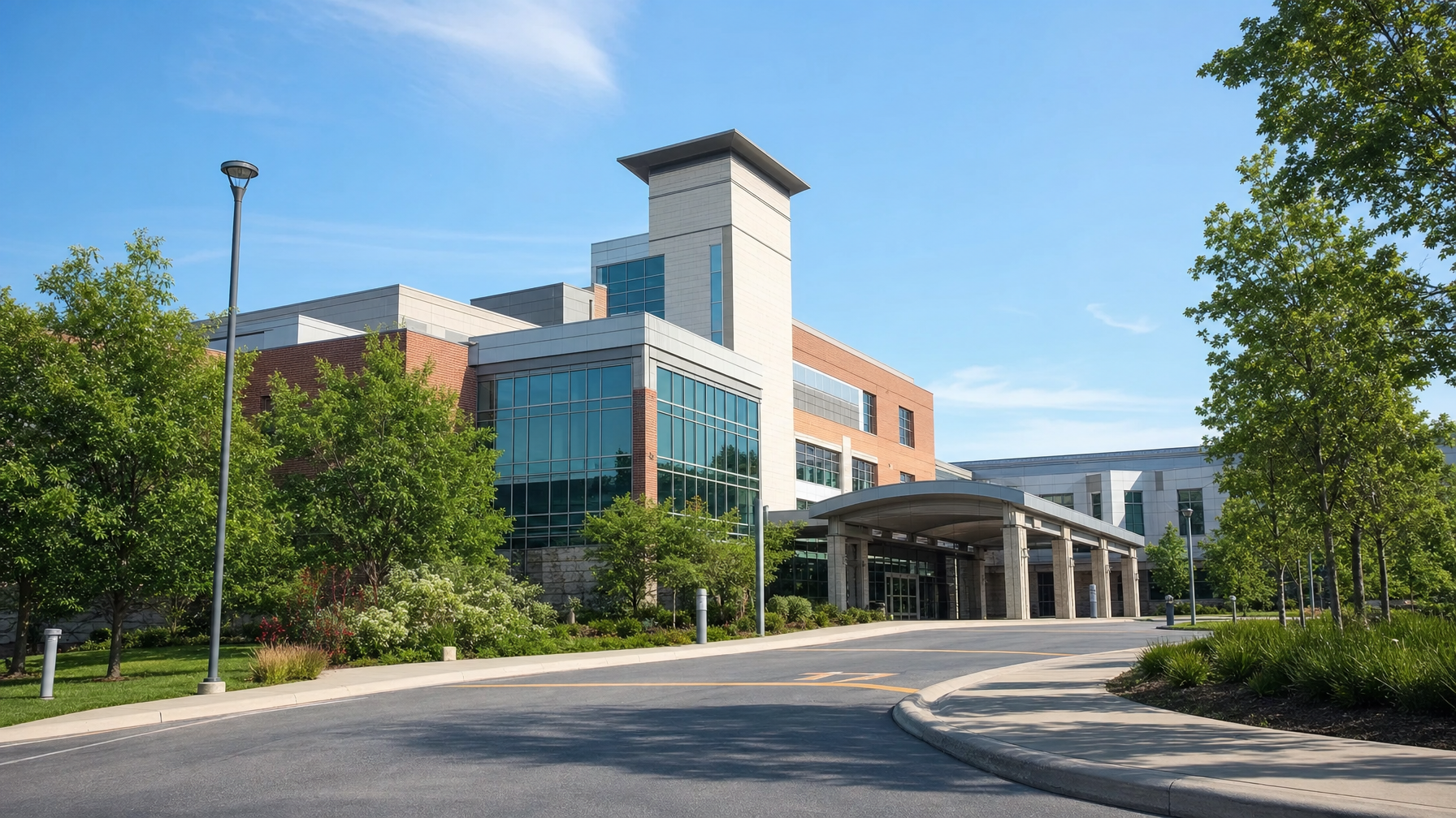 A large modern institutional building with brick, glass, and stone facades stands beside a landscaped driveway and covered entrance under a clear blue sky.